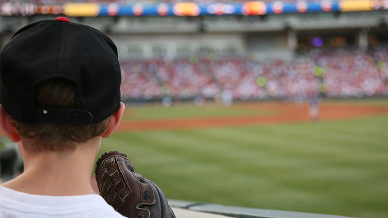 A child in a baseball hat with a baseball glove looks on at a baseball stadium with a game playing.